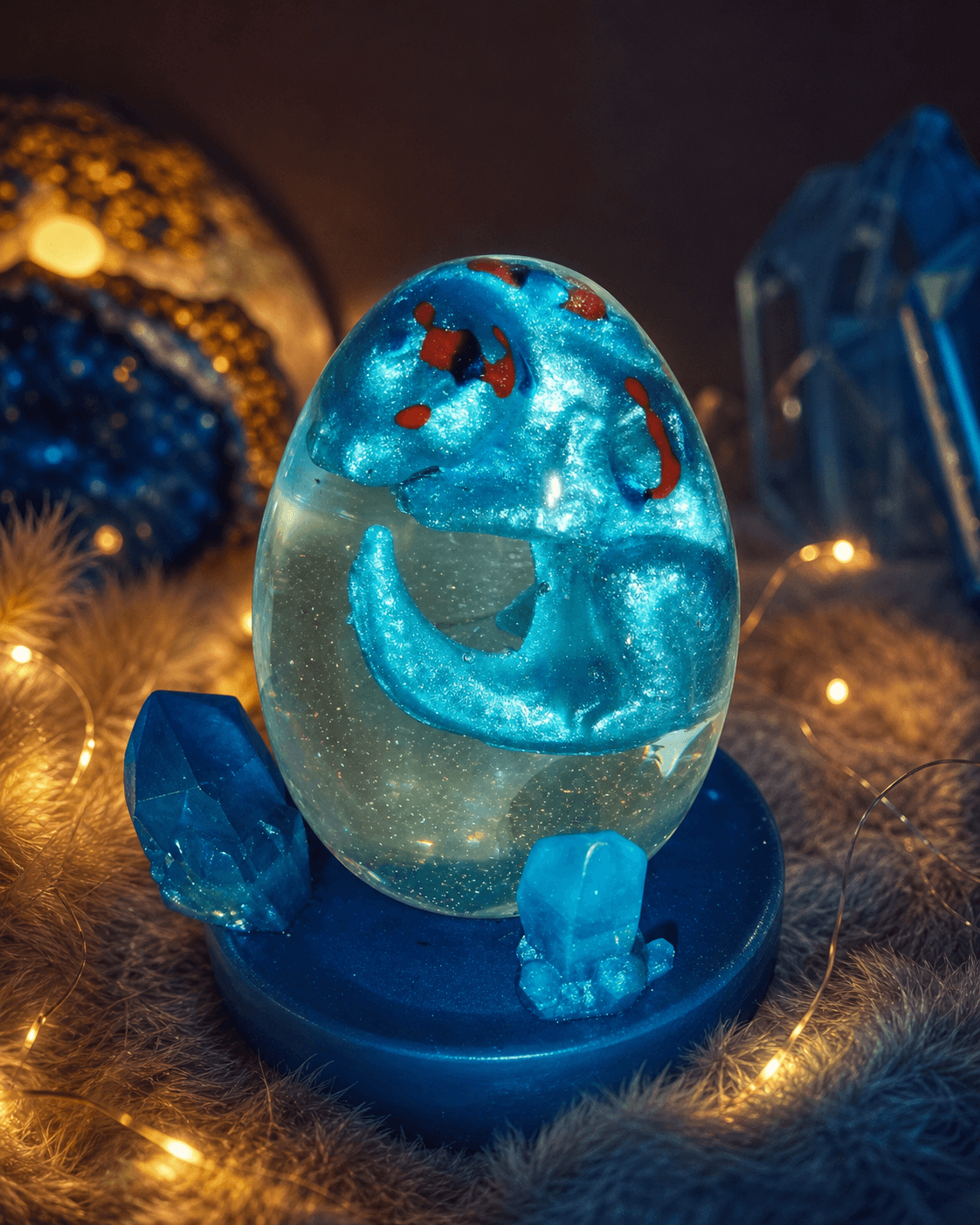 Veilleuse œuf de bébé dragon personnalisé en résine époxy, dragon bleu métallisé et rouge dans son œuf en cristal translucide posé sur un socle bleu avec cristaux de quartz.